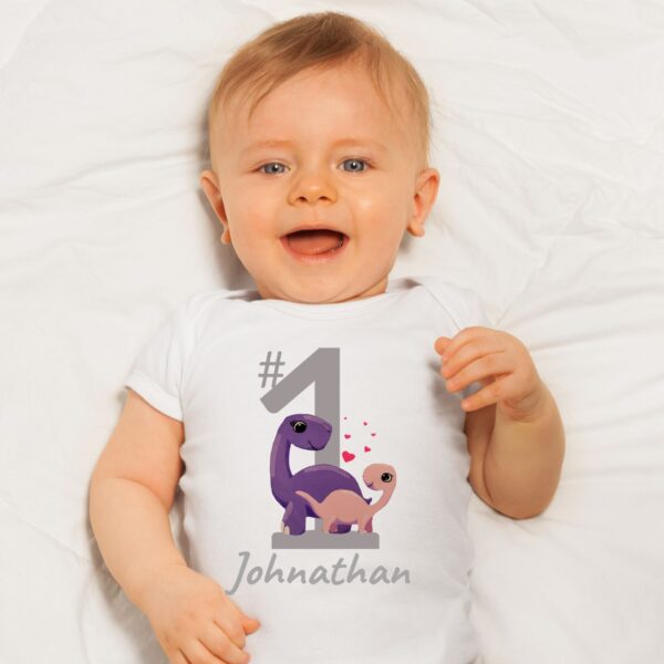 Baby wearing a white summer bodysuit with a dinosaur design, a big number 1 and the baby's name is optional to personalize.