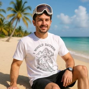 A man sitting on the beach, wearing a white t-shirt with the text "I would rather" and the design of a person skiing.