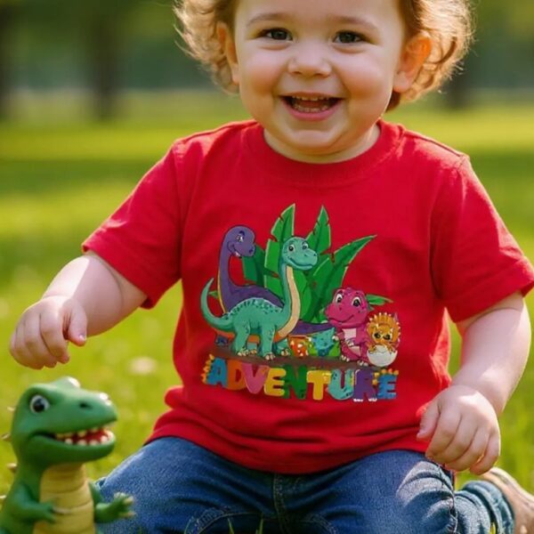 A toddler wearing a red t-shirt with the text: "Adventure" and the design of different colour dinosaurs. The toddler is sitting on the grass and playing with a dinosaur.