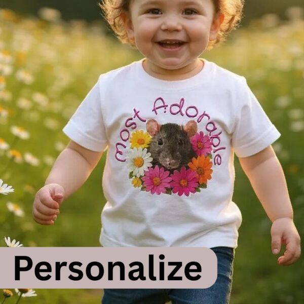 Rat Daisy Personalize Toddler Tshirt Main A toddler wearing a white t-shirt with the text: "Most Adorable" and the design of a cute rat surrounded by colourful daisies. It is optional to personalize the text on the t-shirt. The toddler is walking in a garden full of daisies.