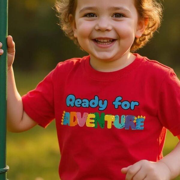A toddler wearing a red t-shirt with the text: "Ready for Adventure". The toddler is playing on a jungle gym.