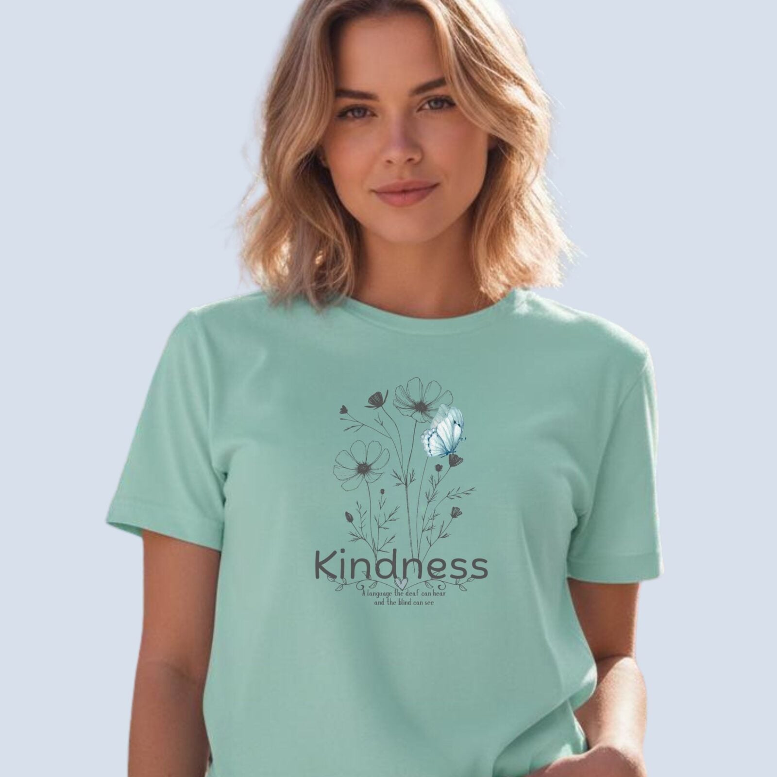 A woman wearing a mint coloured t-shirt with a flower and butterfly design, and the text "Kindness" and "A language the deaf can hear and the blind can see".