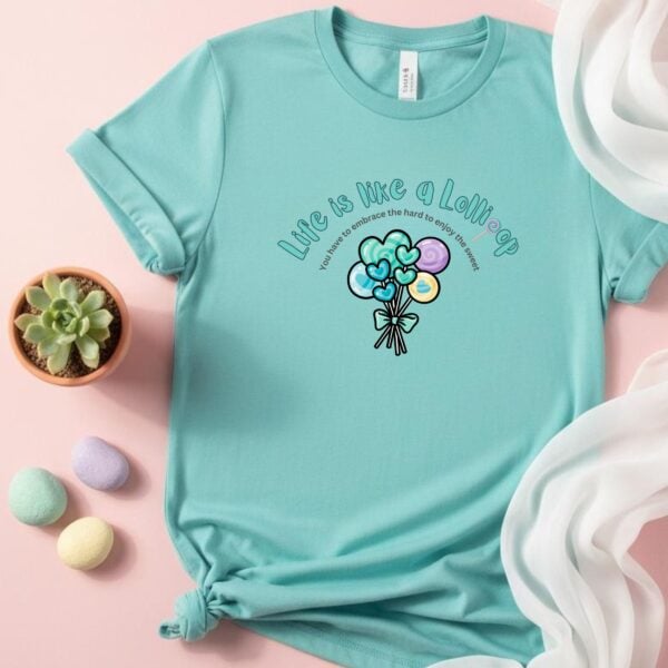 Copy of Flat lay Life is like a Lollipop Tshirt A mint coloured t-shirt with a lollipop design, the text "Life is like a Lollipop" and "you have to embrace the hard to enjoy the sweet". This t-shirt is lying on a pink table next to a pot plant and soft-coloured pebbles.