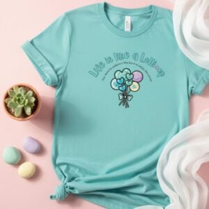 A mint coloured t-shirt with a lollipop design, the text "Life is like a Lollipop" and "you have to embrace the hard to enjoy the sweet". This t-shirt is lying on a pink table next to a pot plant and soft-coloured pebbles.