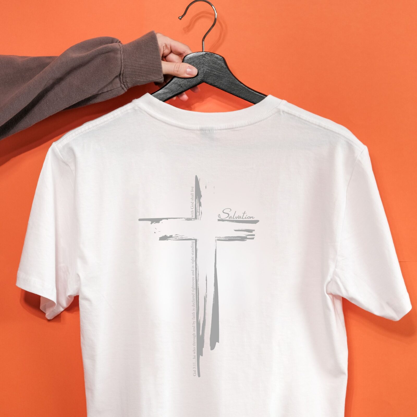 A woman holding a white t-shirt with a cross design on the back of the shirt. This Christian Cross design includes the text "Salvation" and quote Galatians 3:11.