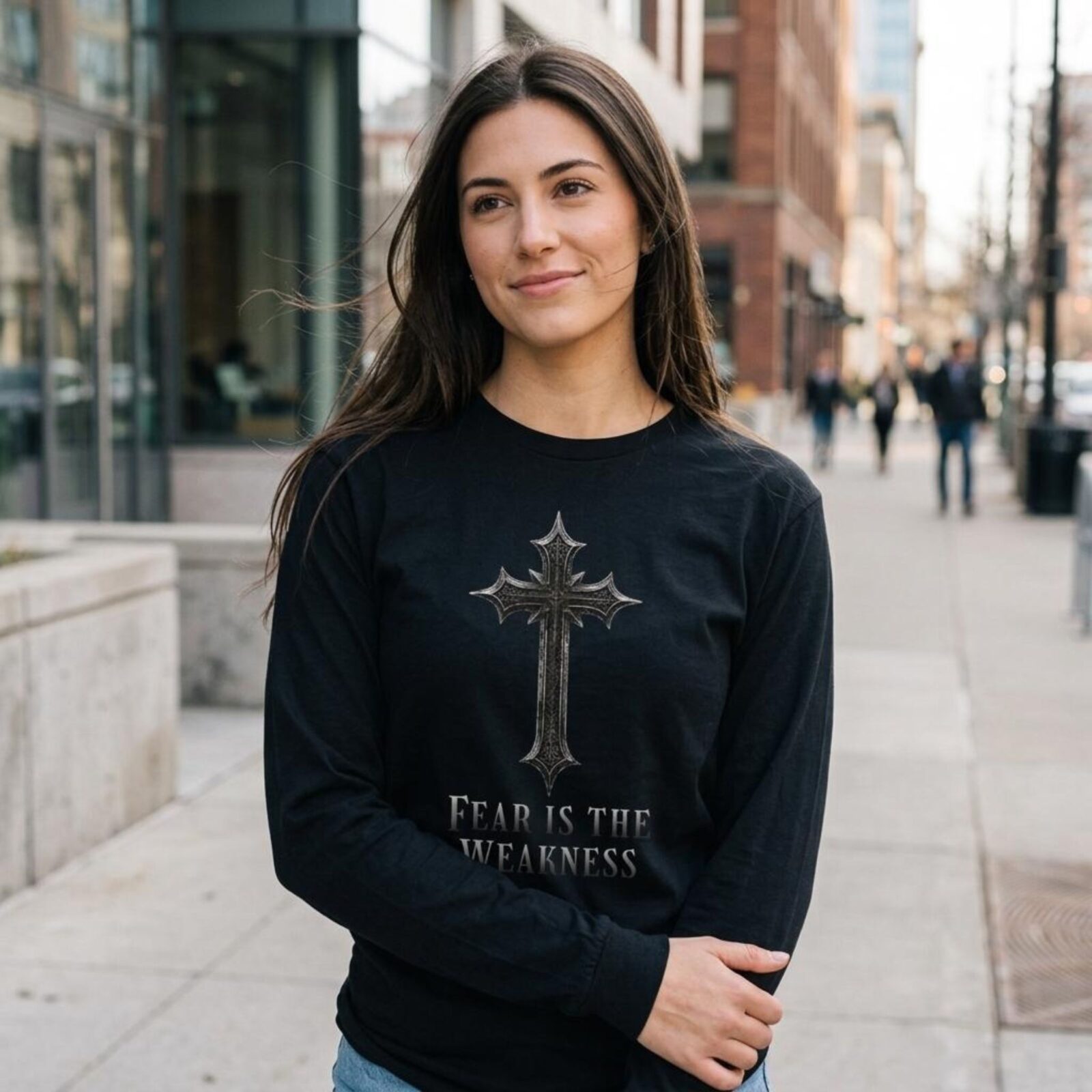A woman wearing a black unisex long sleeve t-shirt with a cross design and the text "Fear is the Weakness".