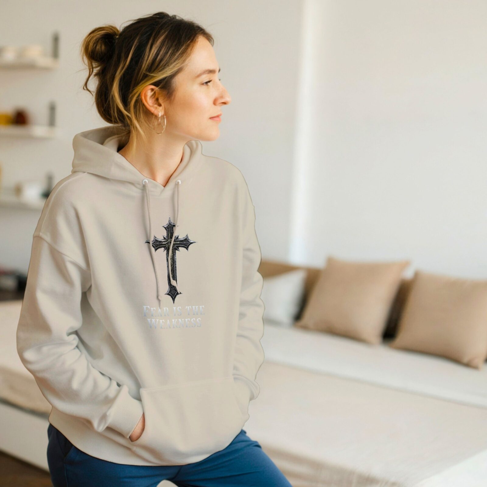 A woman wearing a sand coloured unisex pullover hoodie with a cross design and the text "Fear is the Weakness".