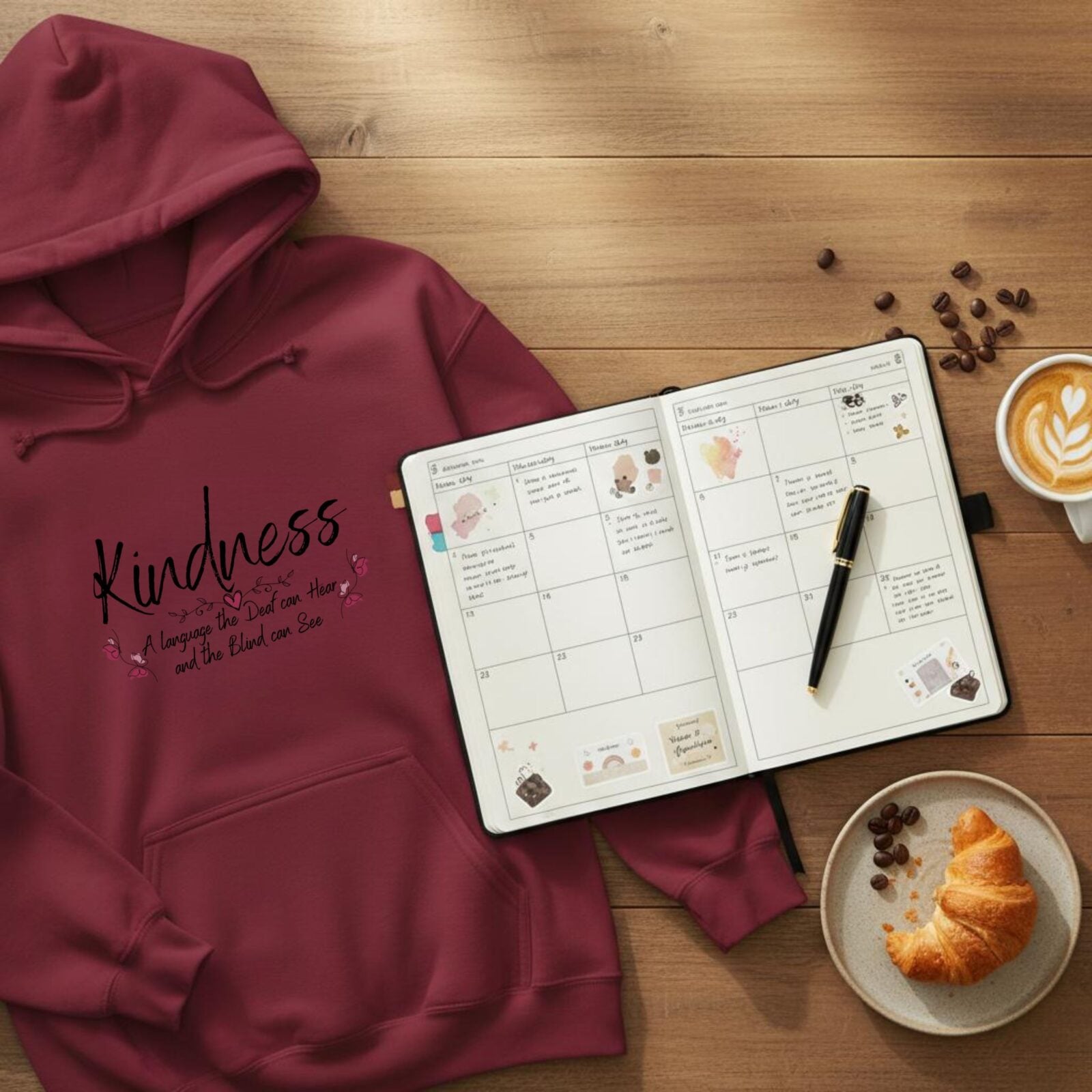 A maroon coloured pullover hoodie with a butterfly design and the text "Kindness, a language the deaf can hear and the blind can see". This hoodie is lying on a table next to a diary and a cup of coffee.