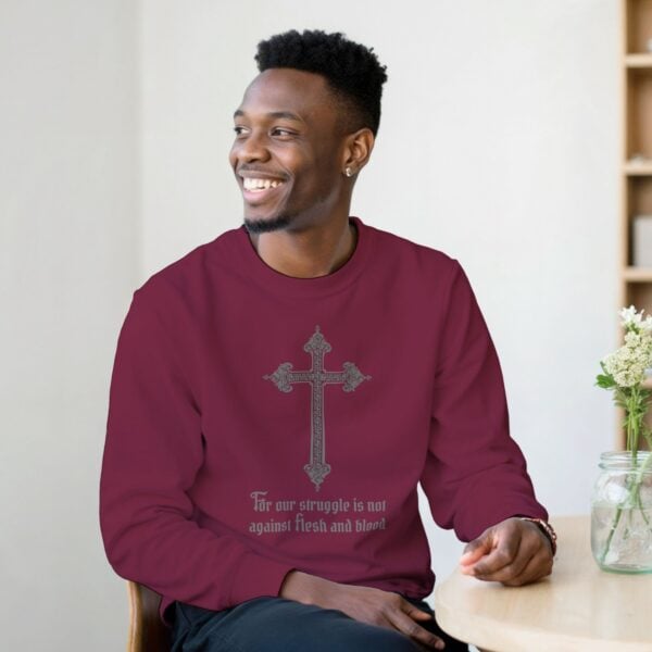 A Man sitting at a table wearing a maroon cross design unisex sweatshirt with the text "For our struggle is not against flesh and blood"