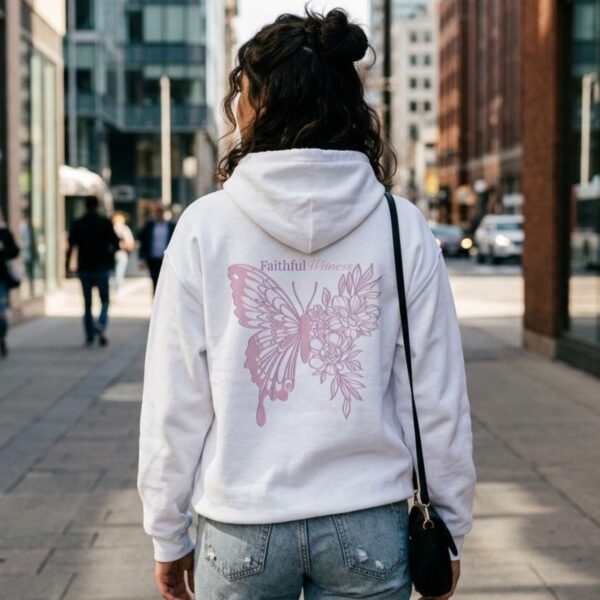 A woman is walking in the city. She is wearing a white pullover hoodie with a butterfly and flower design, and the text "Faithful Witness" printed on the back of the hoodie.