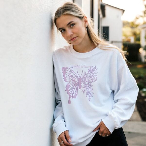 A woman, leaning against a wall, wearing a white sweatshirt with a butterfly and flower design and the text "Faithful Witness" on the front of the sweatshirt.