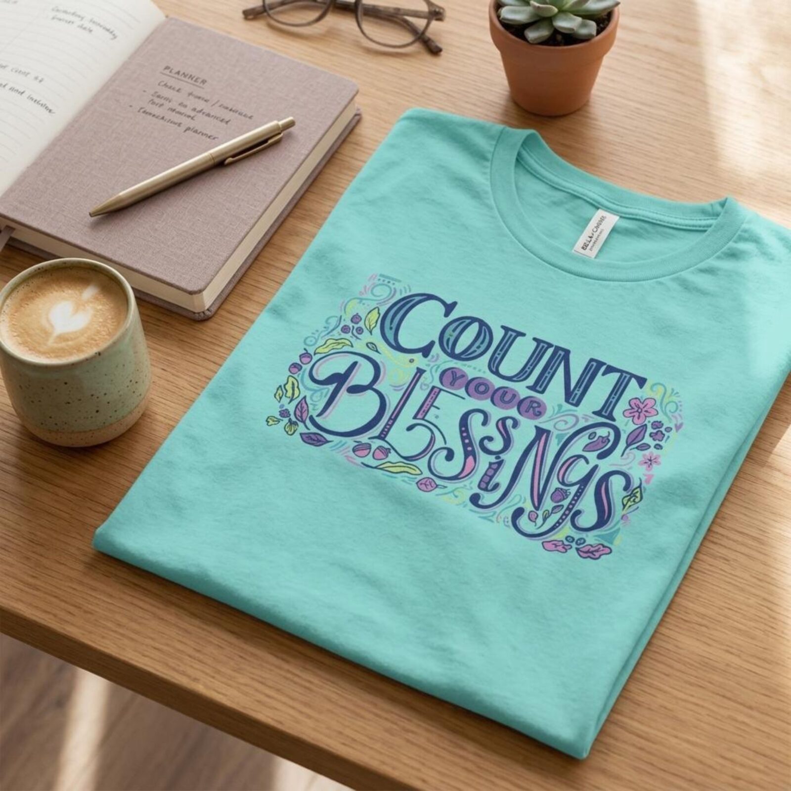 A mint t-shirt with a colourful design and the text "Count your Blessings", neatly folded on a table next to coffee.