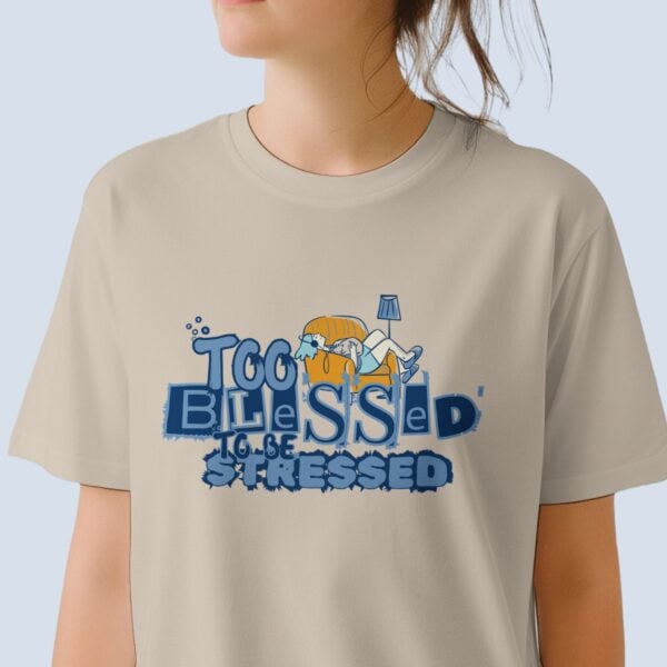 A woman wearing a sand coloured unisex Christian t-shirt featuring the declaration “Too Blessed To Be Stressed” and a graphic of a woman relaxing with her cat, printed on the front of the t-shirt.