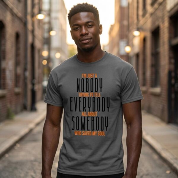 Tell Everybody about Somebody Tshirt Man A man wearing a charcoal coloured unisex Christian t-shirt with the inspirational religious text "I’m just a nobody trying to tell Everybody all about Somebody Who saved my soul". The message celebrates the One who saves and encourages us to tell others about Jesus.